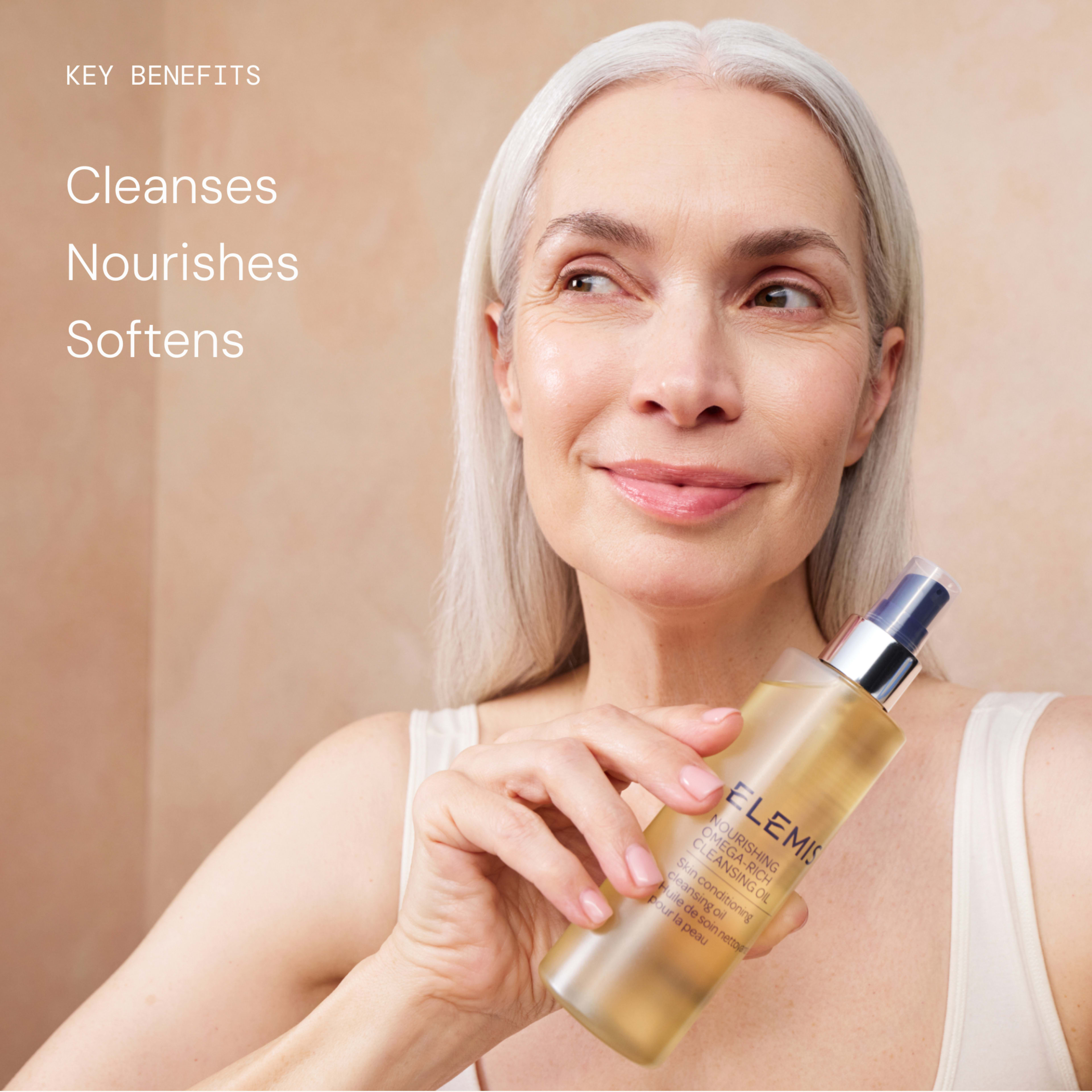 Smiling mature woman with long grey hair holding an ELEMIS facial oil bottle near her face against a neutral background. Text reads: “Key benefits: cleanses, nourishes, softens.”