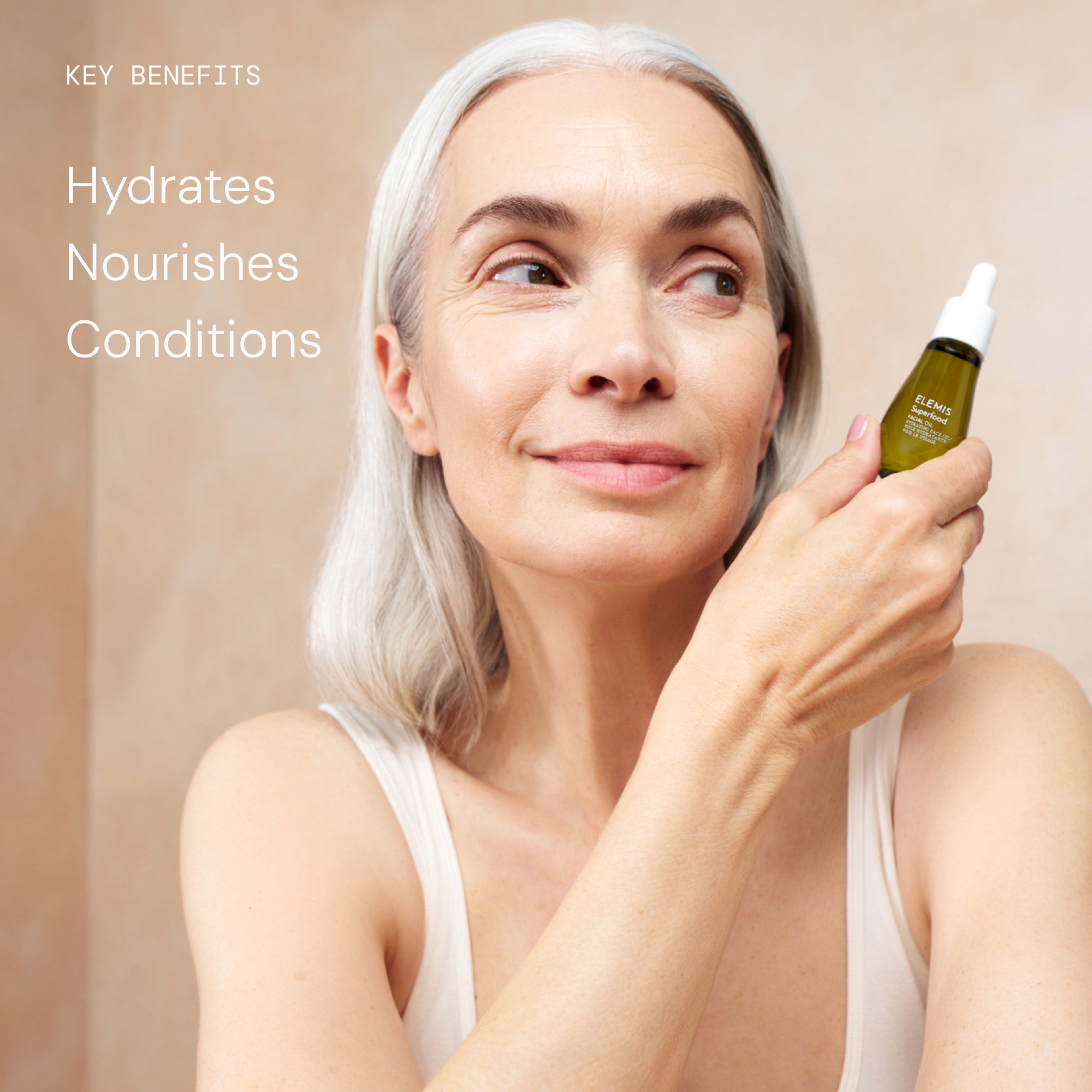 Smiling mature woman with grey hair holds an ELEMIS facial serum dropper bottle to her cheek against a neutral background. On the left, text reads: “Key benefits Hydrates Nourishes Conditions”.