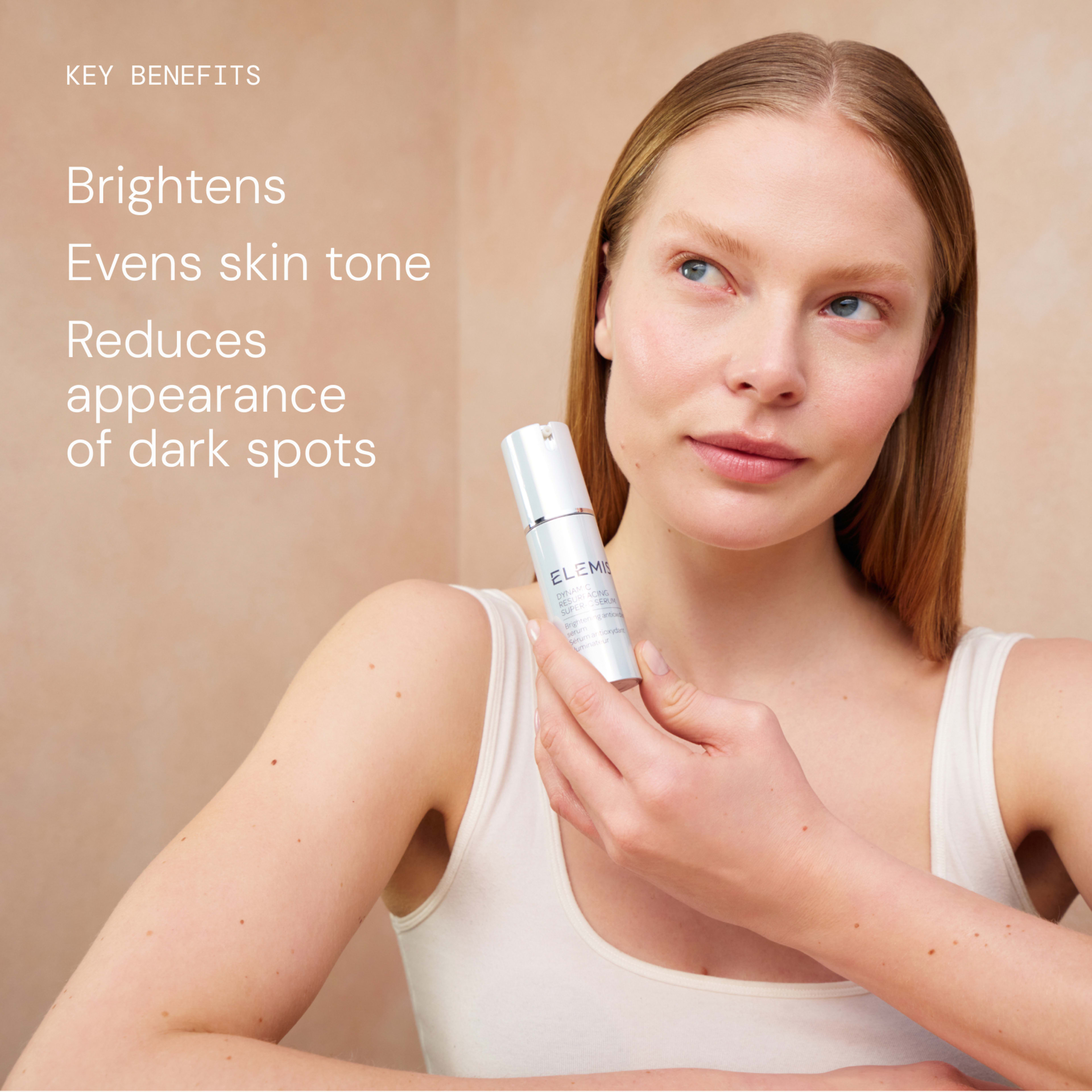 Woman holding an ELEMIS skincare serum bottle to her cheek, looking upwards, against a neutral background. On the left, text reads: “Key benefits: Brightens, Evens skin tone, Reduces appearance of dark spots.”