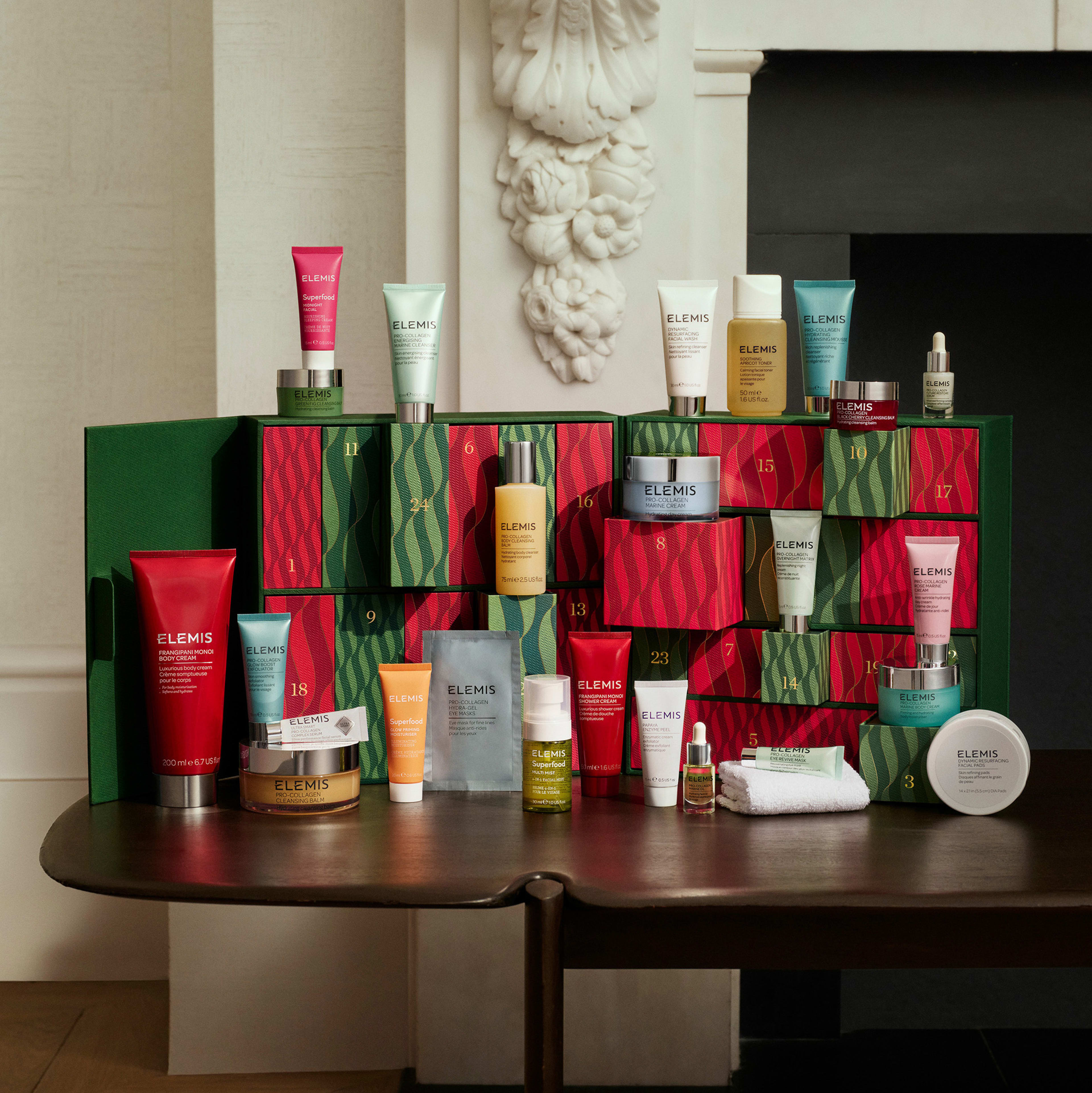 ELEMIS holiday advent calendar displayed on a wooden table, with numbered red and green drawers surrounded by assorted skincare products including cleansers, moisturisers, oils and creams in colourful tubes, jars and bottles arranged in front of a fireplace.