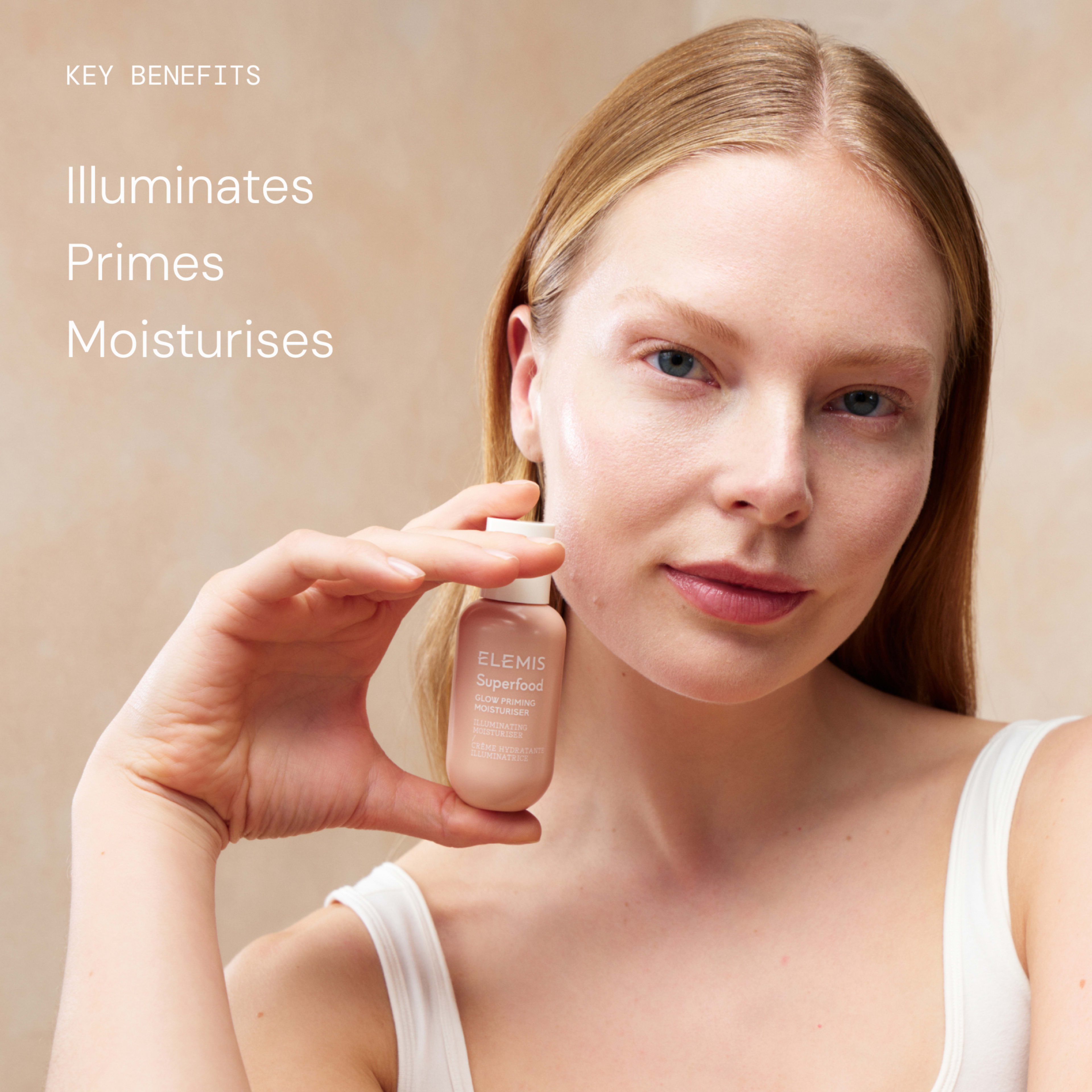 Woman with dewy skin holding a bottle of ELEMIS Superfood Glow Priming Moisturiser beside her face against a neutral background. On the left, text reads: “Key benefits: Illuminates, Primes, Moisturises.”