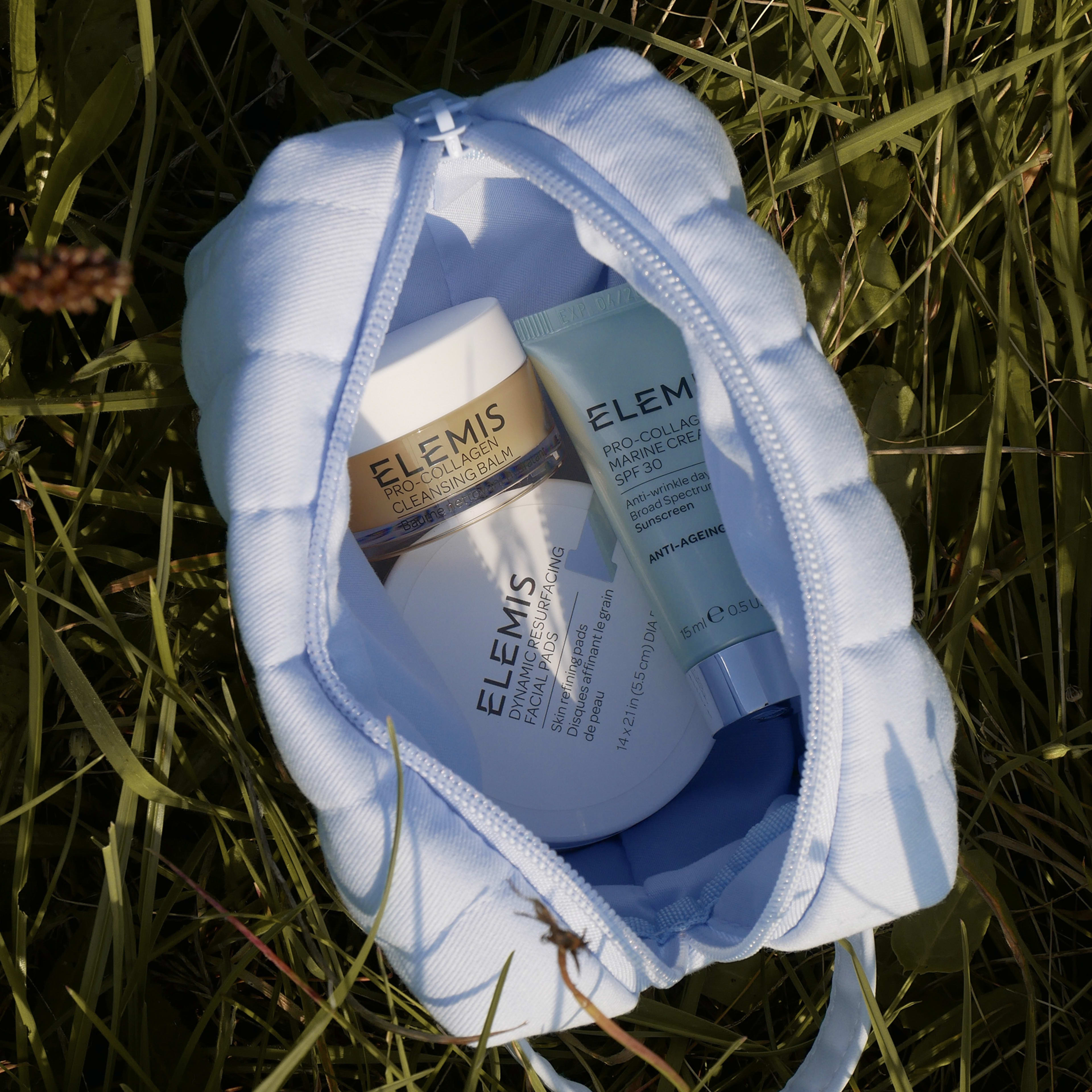 Open pale blue pouch lying in grass, holding three ELEMIS skincare minis: Pro-Collagen Cleansing Balm, Pro-Collagen Marine Cream and Pro-Collagen Marine SPF30, with product labels partly visible in natural sunlight.