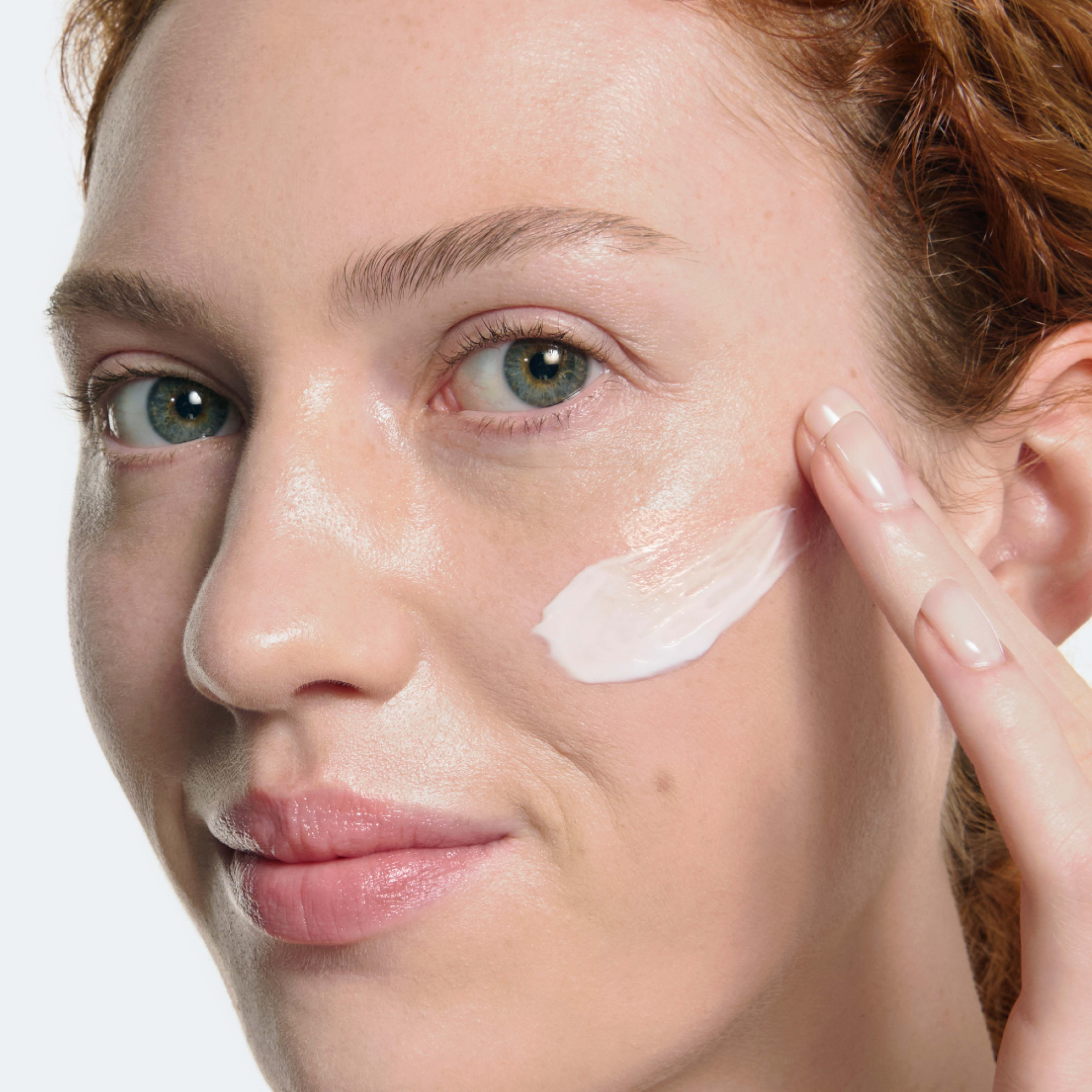 Woman applying a swatch of white ELEMIS skincare cream to her cheek with her fingertips, showing how to use the product on clean, hydrated skin.