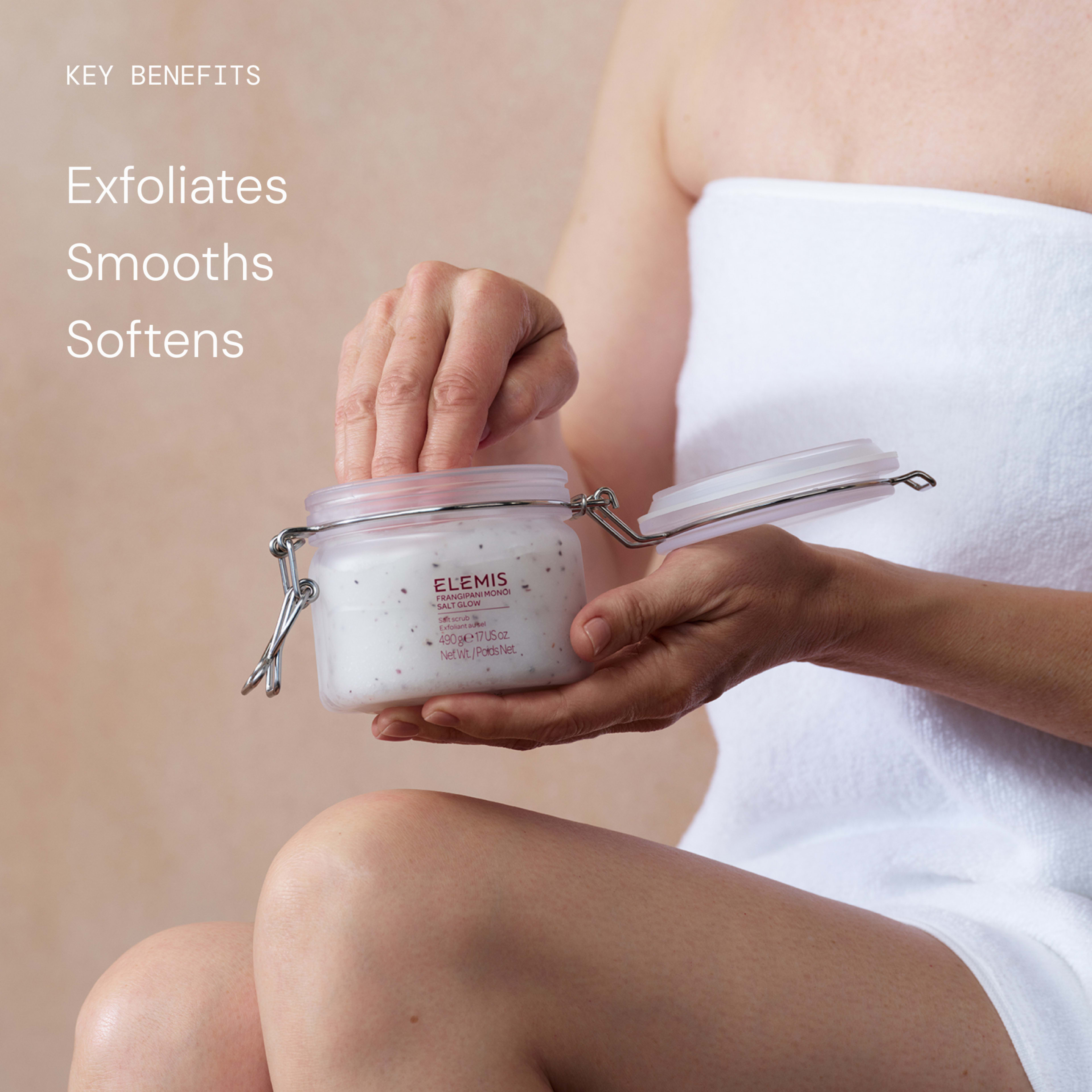Person in a white towel scooping body scrub from a jar of ELEMIS Frangipani Monoi Salt Glow, 490g, against a neutral background. On the left, key benefits text reads: “Exfoliates, Smooths, Softens”.
