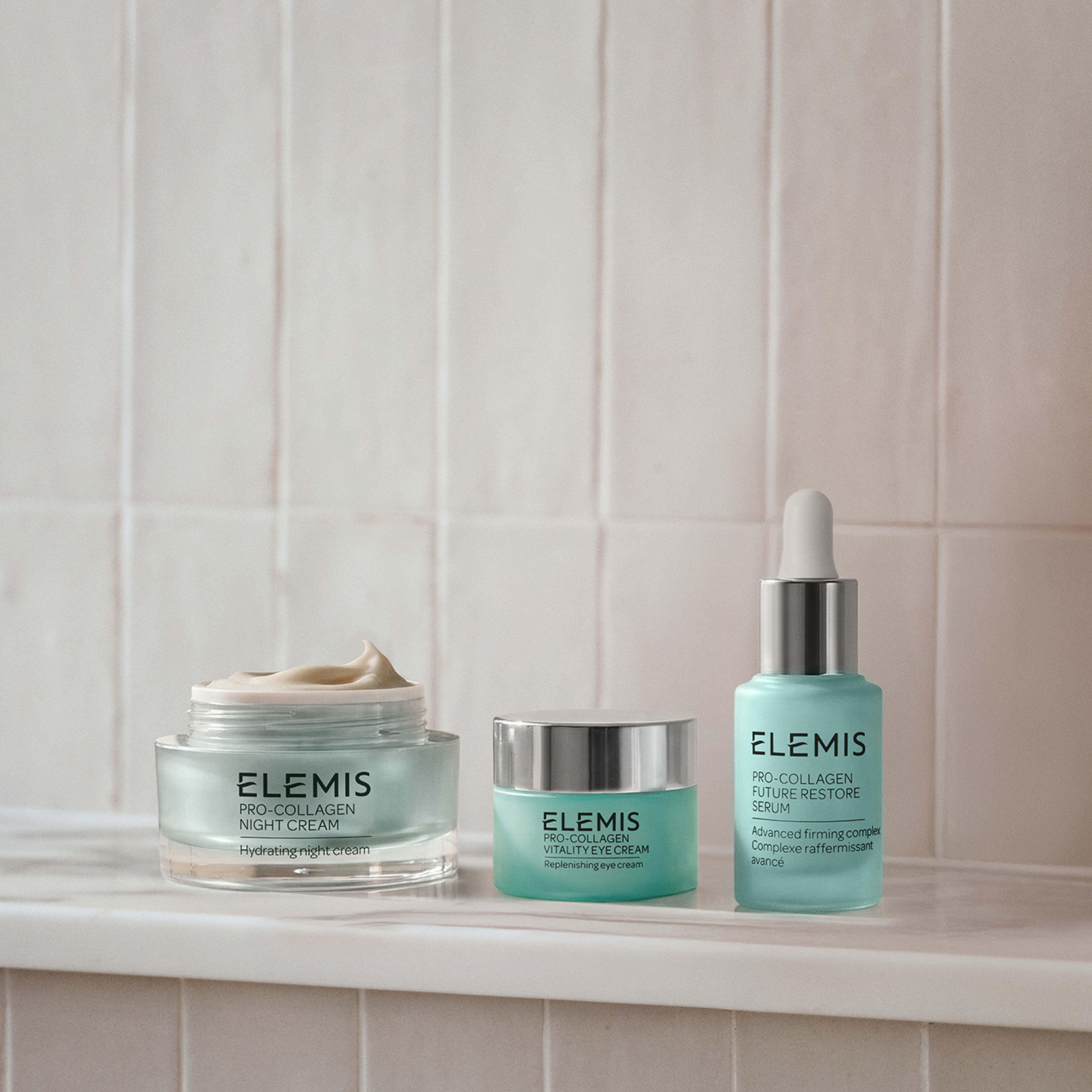 ELEMIS Pro-Collagen skincare trio on a bathroom shelf: open Night Cream jar showing cream, Vital Eye Cream jar with silver lid, and Future Restore Serum bottle with dropper against pale tiled wall.