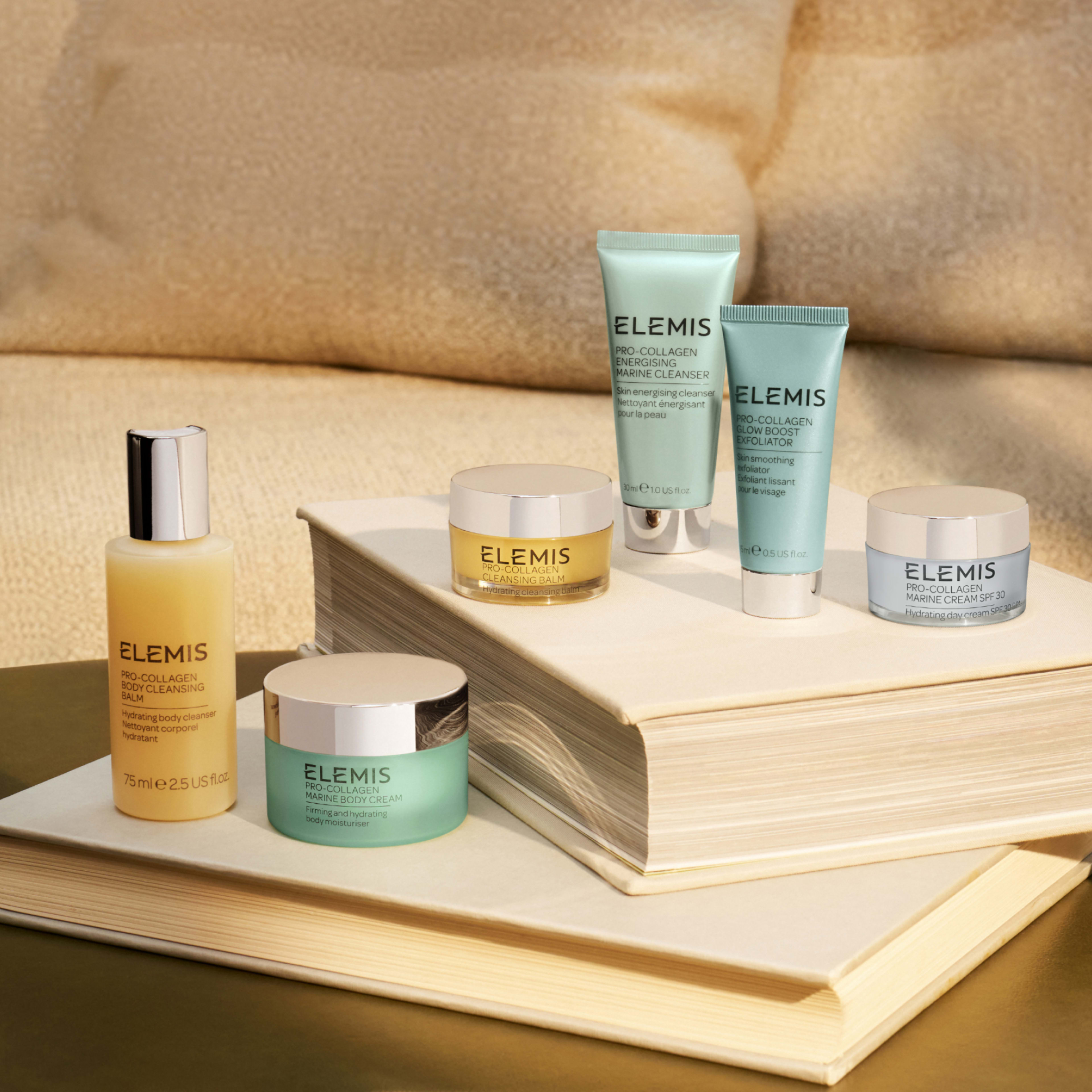 Collection of ELEMIS Pro-Collagen skincare products, including cleansers, creams and balms in pastel tubes and jars, arranged on two stacked books on a neutral sofa, styled in warm natural light.
