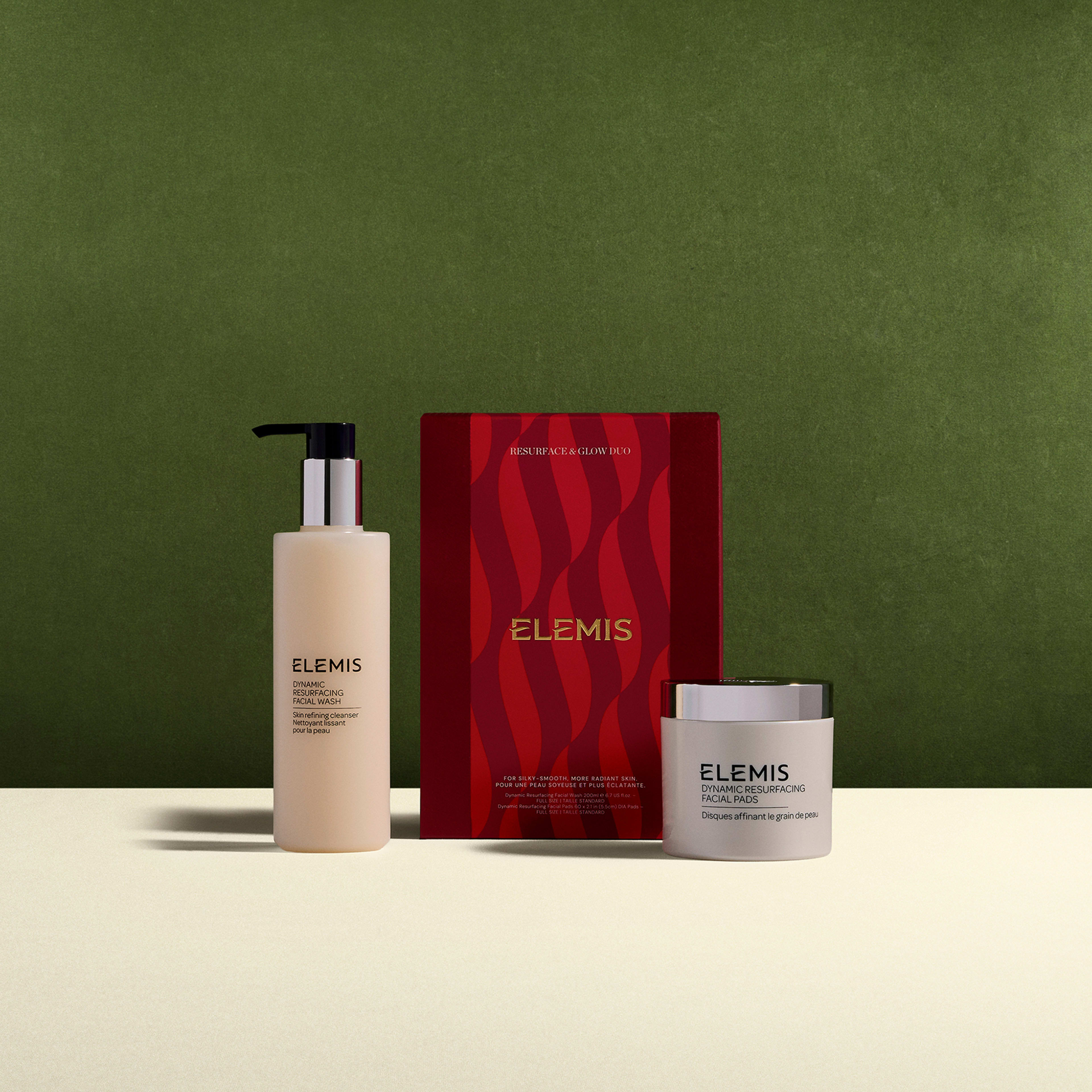 ELEMIS Dynamic Resurfacing skincare set with pump bottle facial wash, tub of facial pads and red gift box labelled “ELEMIS Resurface & Glow Duo”, displayed on a cream surface against a green background.