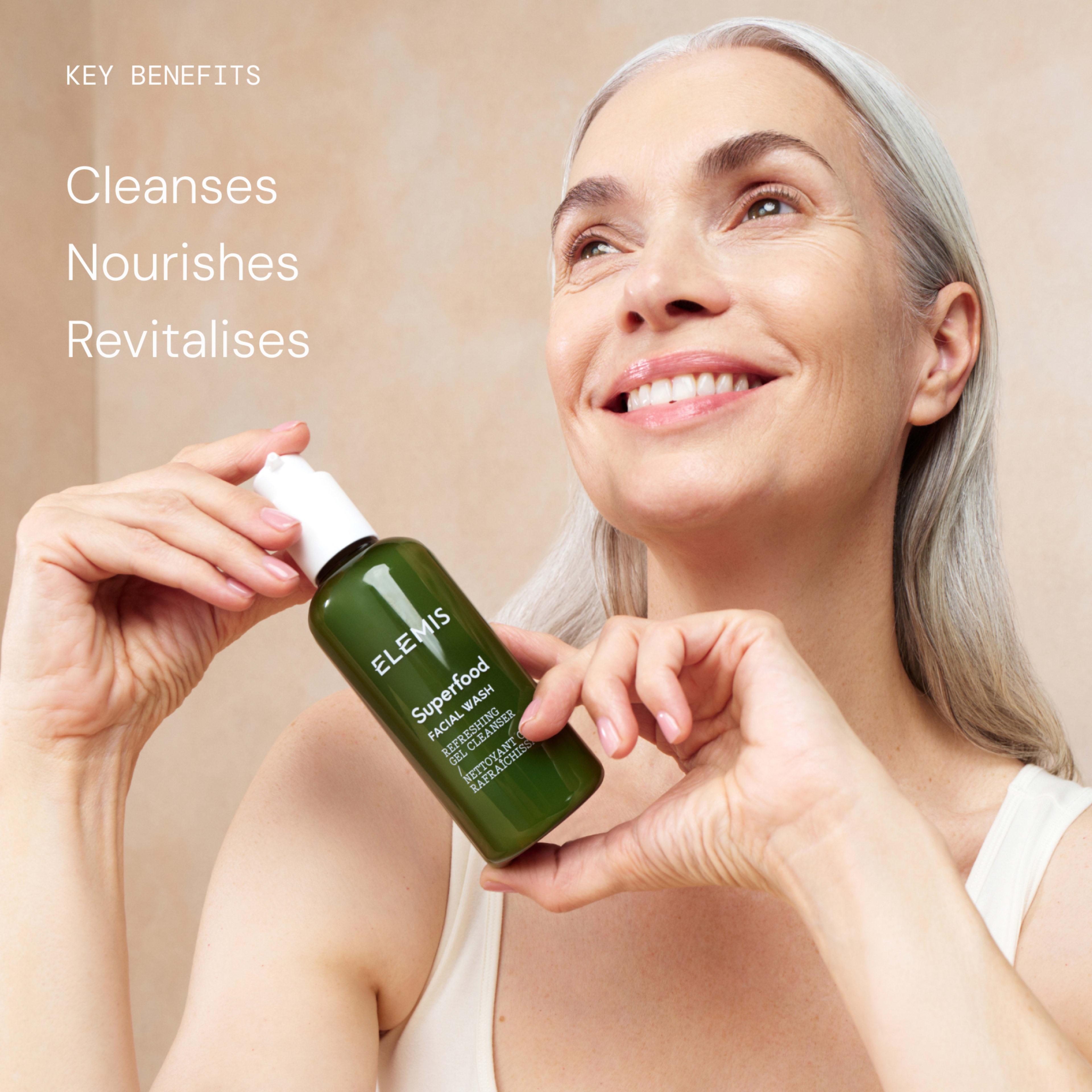 Smiling mature woman with grey hair holding a green bottle of ELEMIS Superfood Facial Wash towards the camera. On the left, text reads: “Key benefits: Cleanses, Nourishes, Revitalises.”