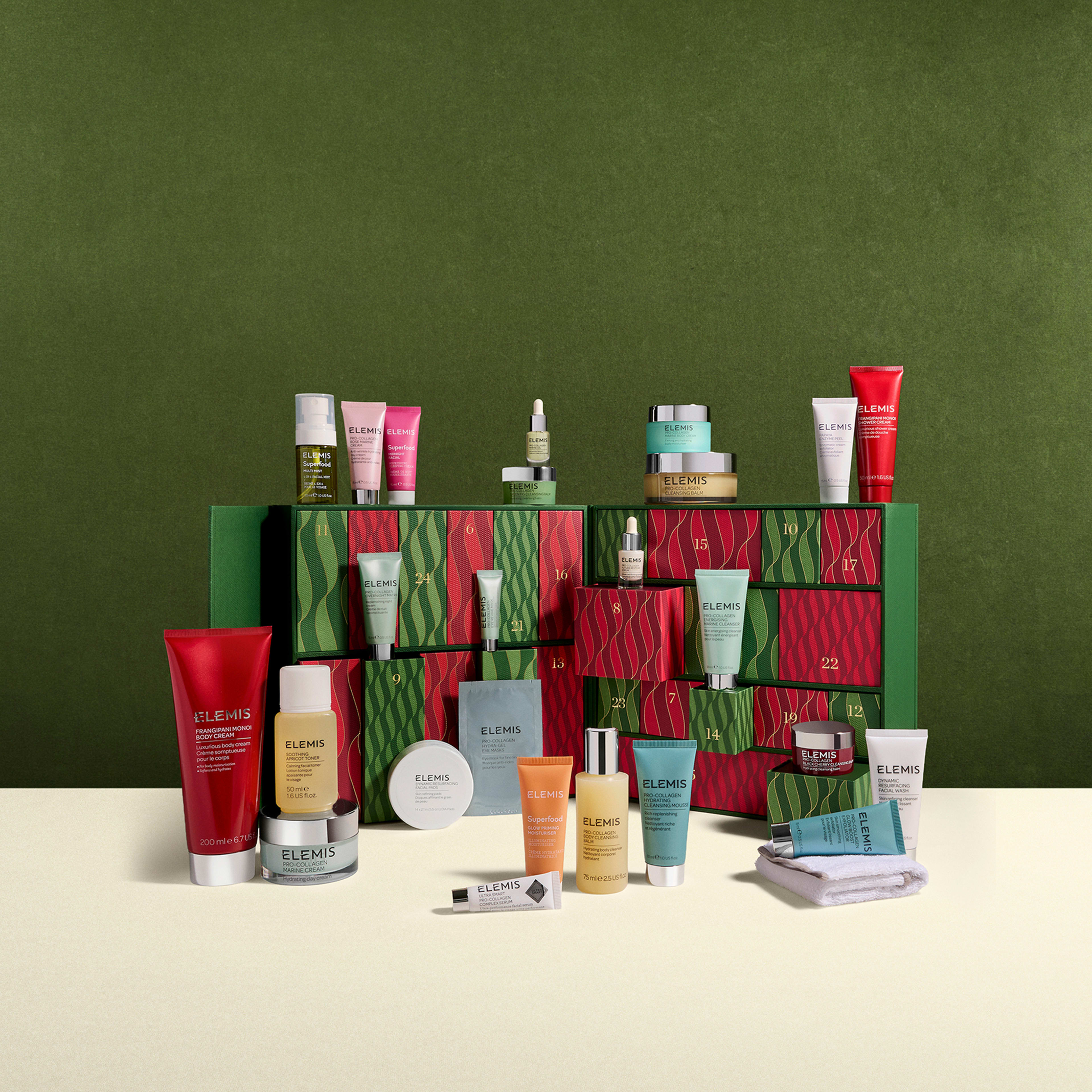 ELEMIS Christmas advent calendar in green and red with numbered drawers, surrounded by an assortment of ELEMIS skincare products including cleansers, moisturisers, serums and balms arranged neatly in front on a cream surface against a green background.