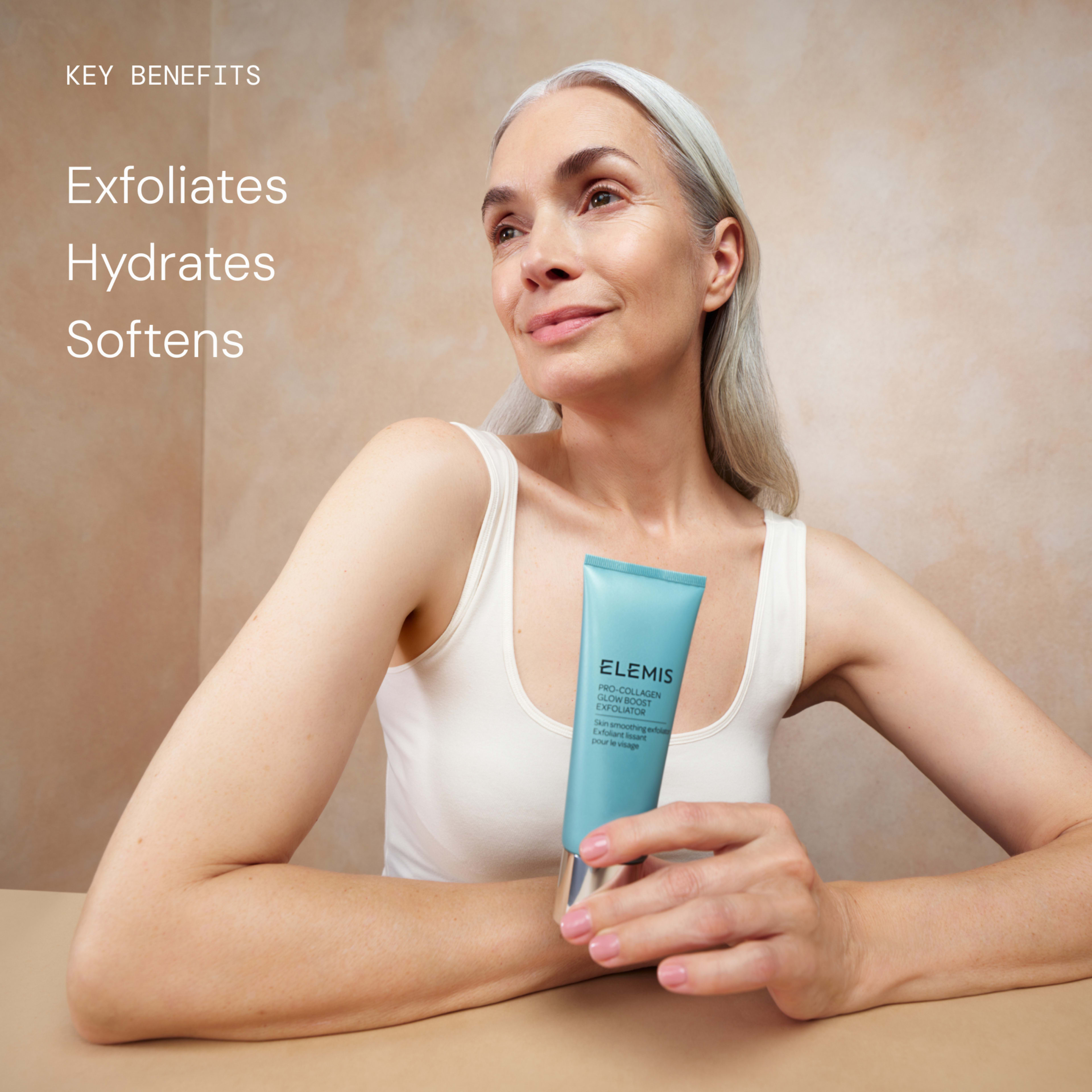 Smiling woman with long grey hair in a cream vest holding a turquoise tube of ELEMIS Pro-Collagen Glow Exfoliator against a neutral background. On the left, text reads: “Key benefits: Exfoliates, Hydrates, Softens.”