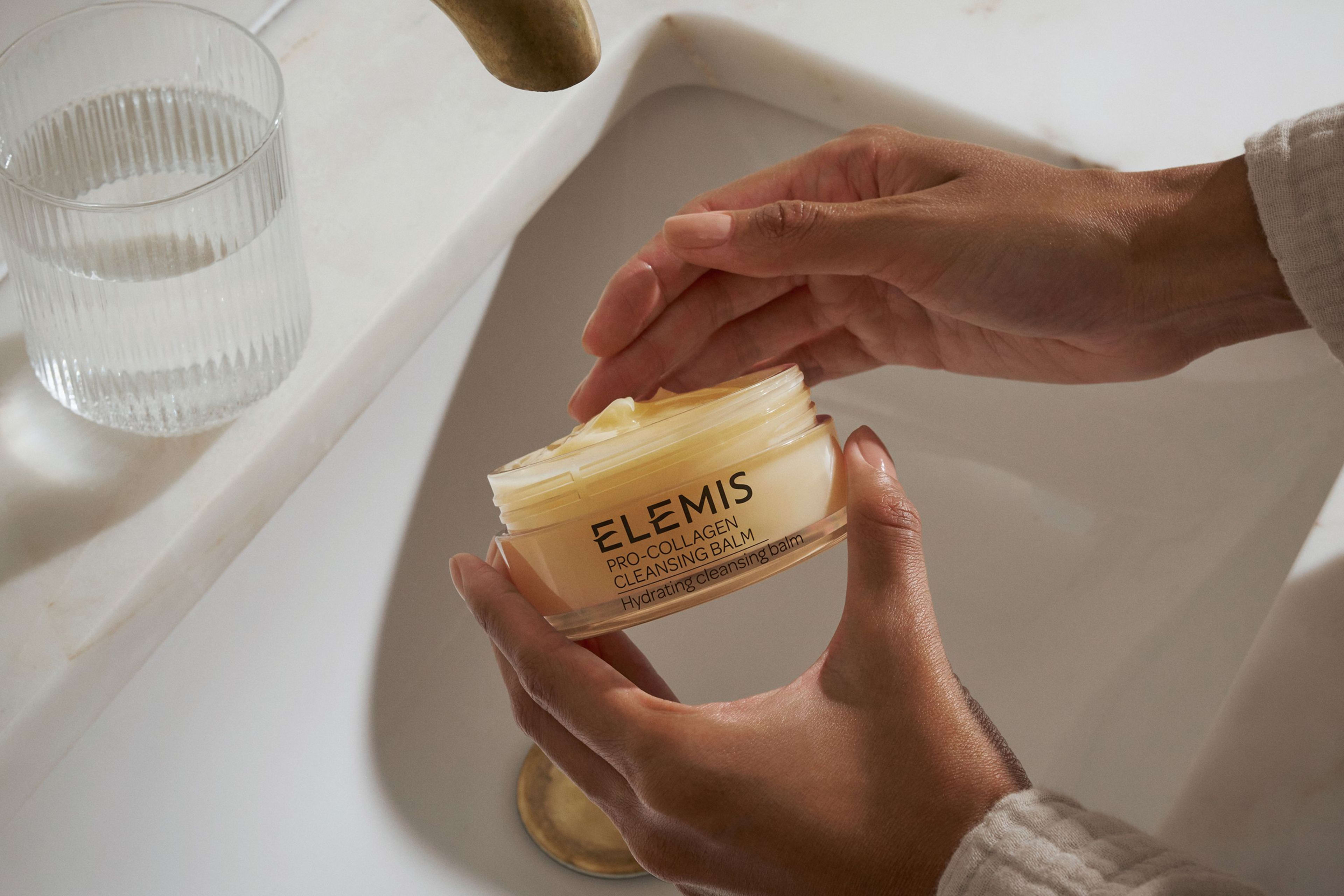 A hand holds three stacked Elemis Pro-Collagen Cleansing Balms (green, yellow, pink) against a bright blue sky and white flowers.