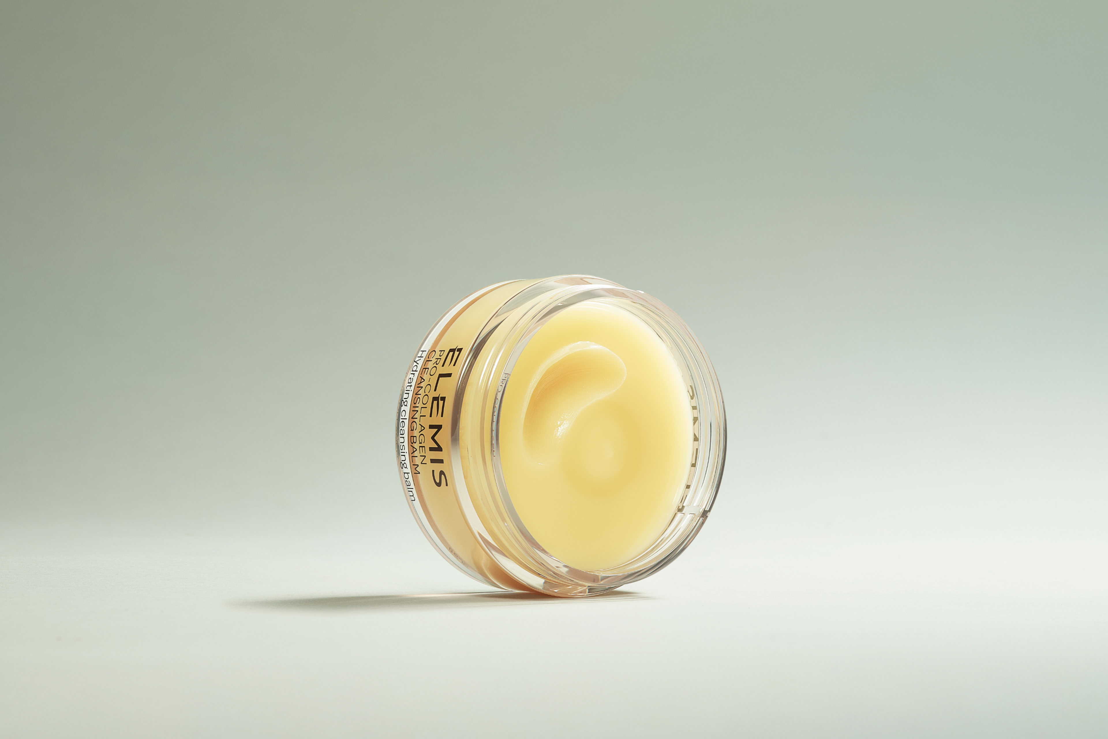 Two Elemis Pro-Collagen skincare products, a mint green cleanser bottle and a yellow cleansing balm jar, sit in shallow water against a dark blue background.